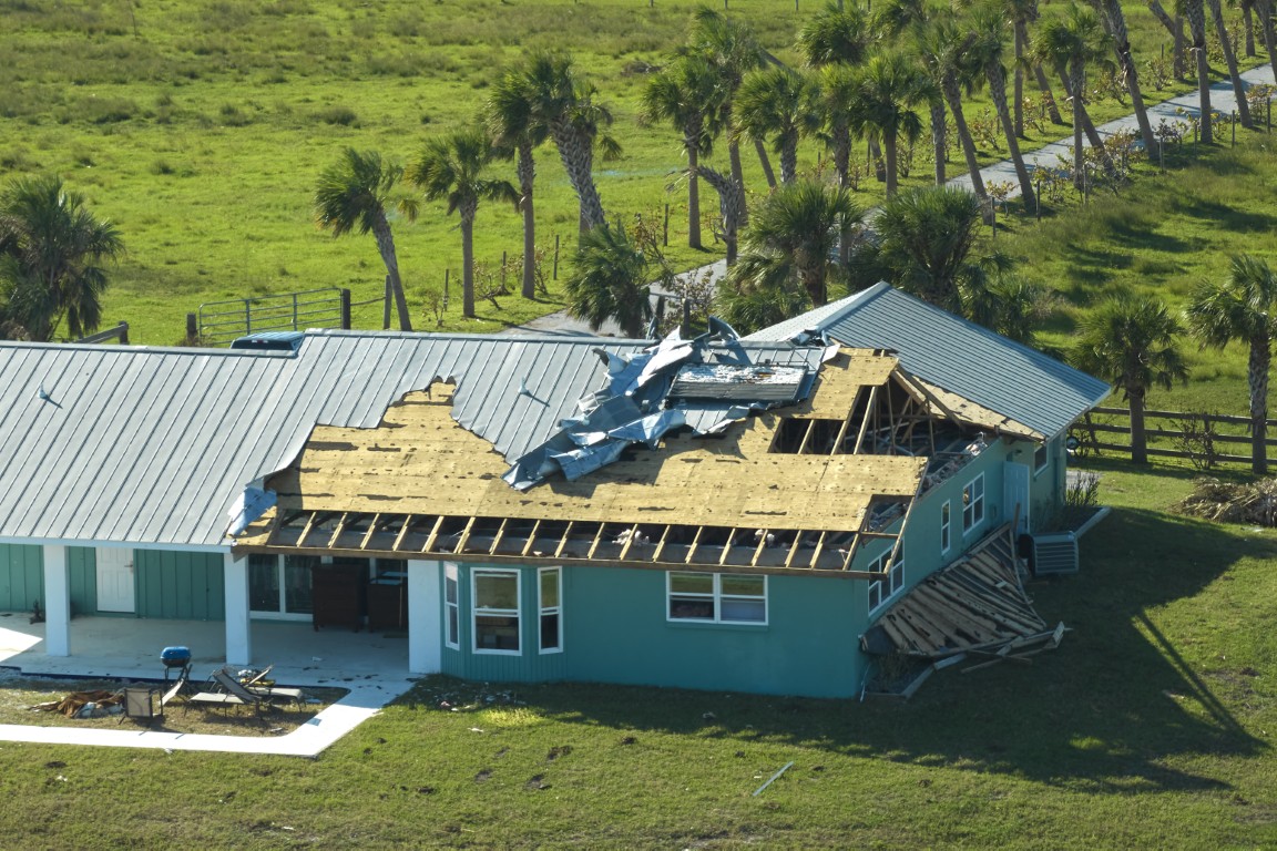 Turquoise single-story house with a severely collapsed roof, exposed wooden framing and scattered debris, set on a green lawn with palm trees in the background.